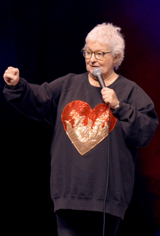 Janey Godley stands on stage holding a microphone. She has short white hair, glasses and wears a great sweatshirt with a red and gold glittery heart on it