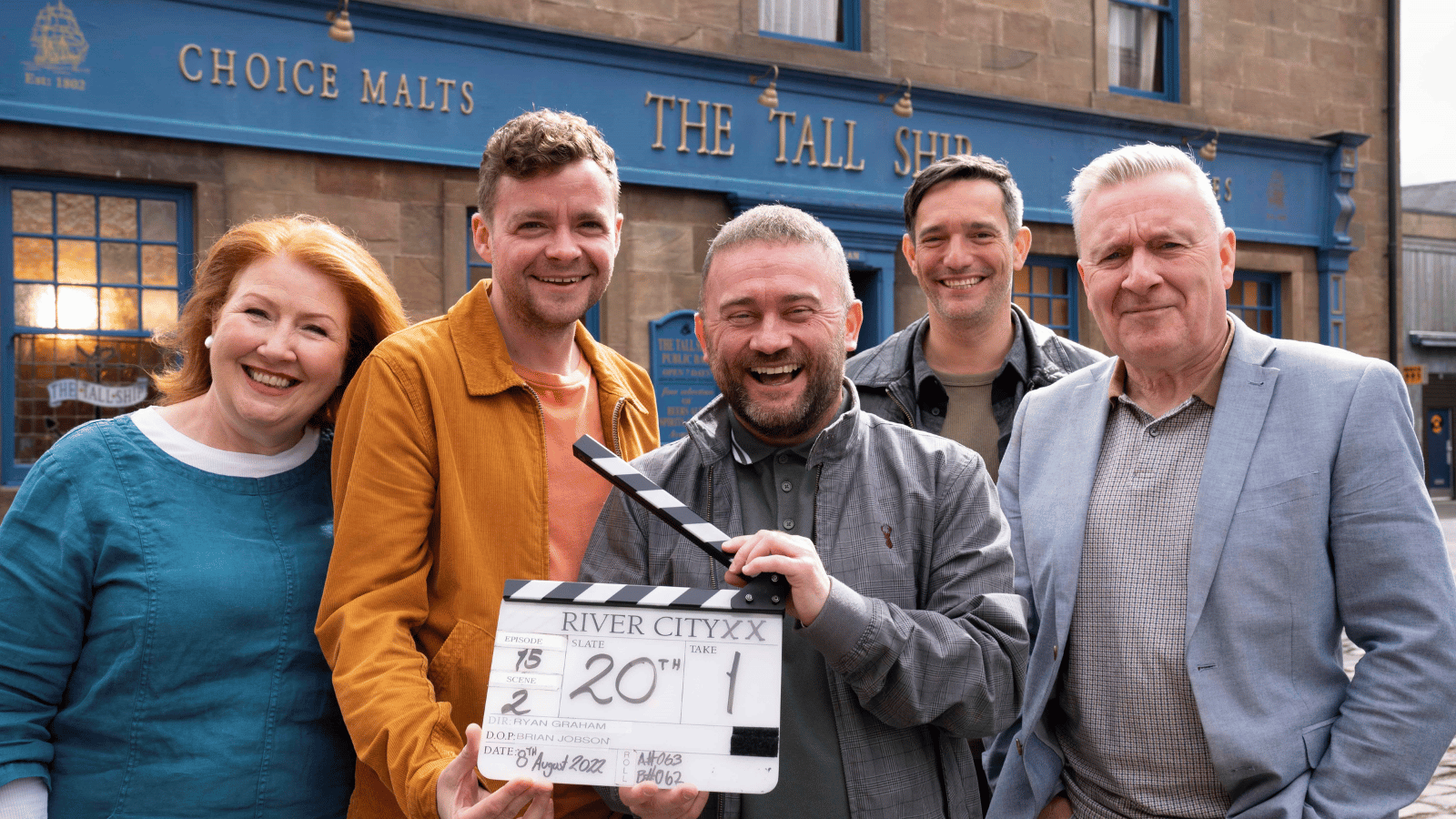 Cast of River City's 20th Episode celebrations standing on set, holding a clapper board.