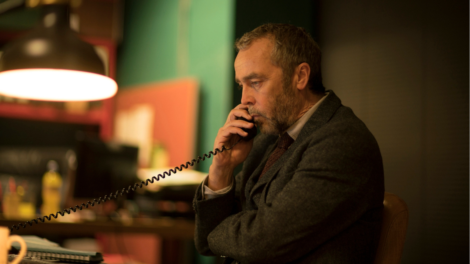 John Hannah sits in a dimly lit room with a telephone to his ear in The Victim